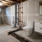 adding bathroom to basement plumbing cost: Basement bathroom under construction with exposed drain pipes and a new white vanity